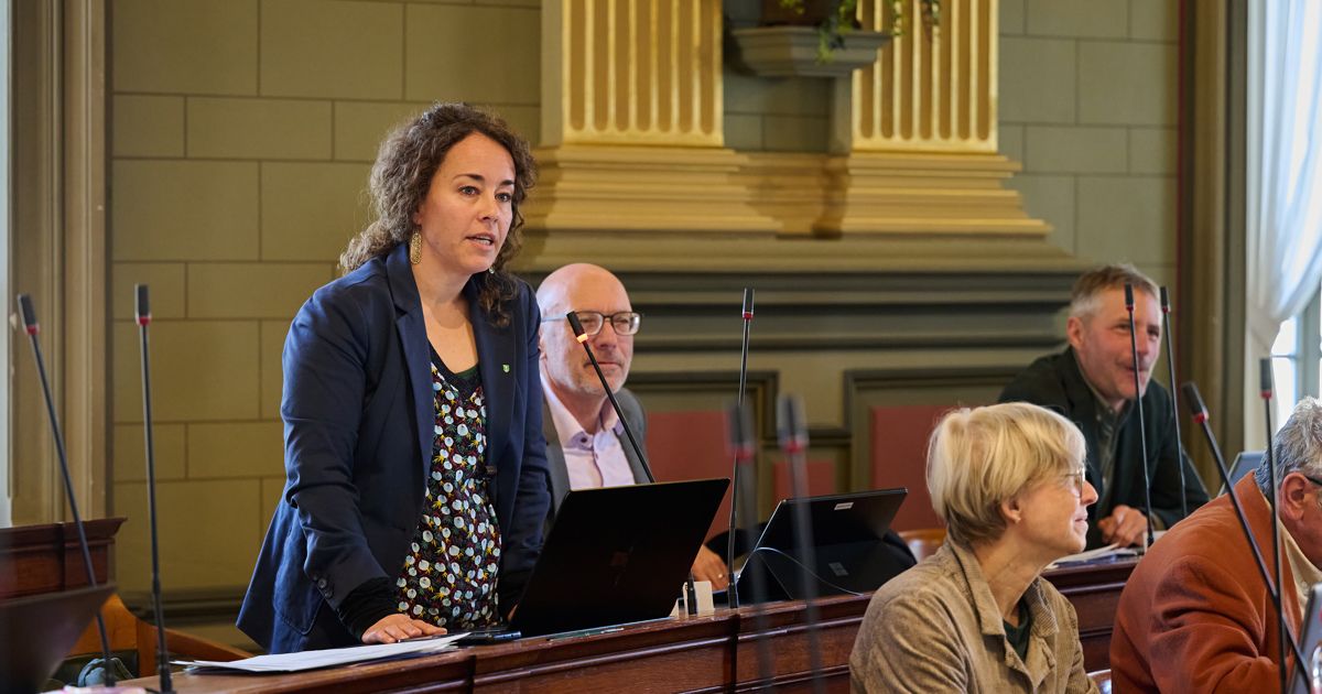 Eine Frau mit lockigem Haar steht an einem Rednerpult und spricht in ein Mikrofon. Hinter ihr lauscht ein Mann mit Brille. Vor ihr stehen zwei Laptops auf dem Tisch. Eine andere Frau sitzt in der Nähe und schaut zu.