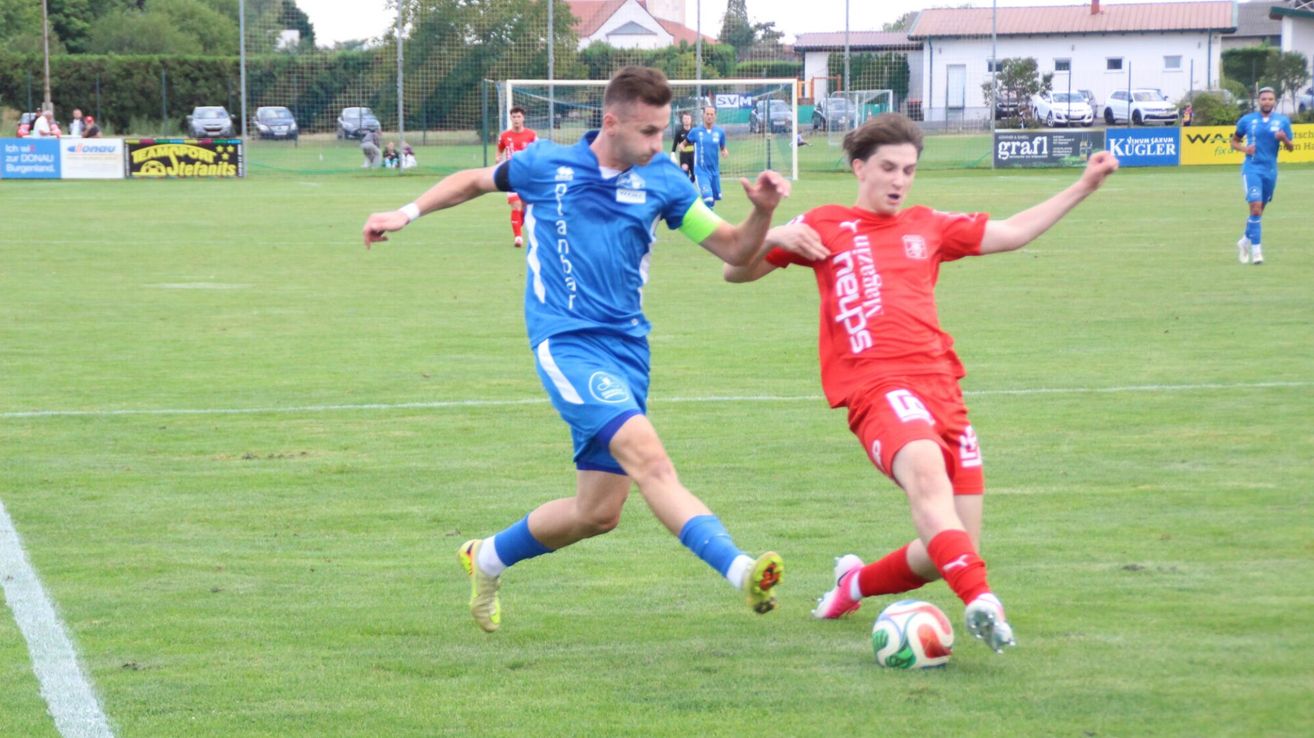 Zwei Fußballspieler, einer in Blau und einer in Rot, befinden sich mitten im Spiel auf einem Rasenfeld. Der Spieler in Blau versucht, dem Spieler in Rot den Ball zu stehlen.