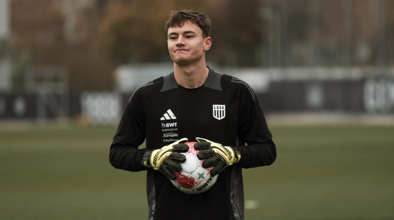 Ein Fußballspieler in einem schwarzen Trikot mit weißen Akzenten hält einen Fußball in beiden Händen. Das Trikot hat mehrere Logos, darunter 'LASK' und 'BWT Energie AG.' Der Spieler steht auf einem Rasenfeld.