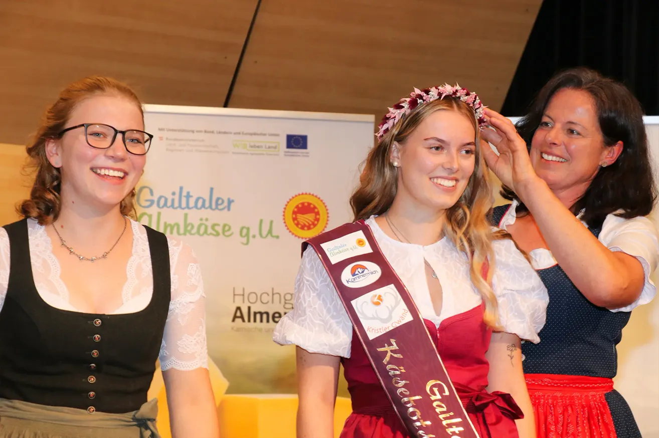 Eine Frau mit einem roten Kleid und einer Blumenkrone lächelt und posiert für ein Foto mit zwei anderen Frauen. Hinter ihr befindet sich ein weißer Banner mit Text und Logos.