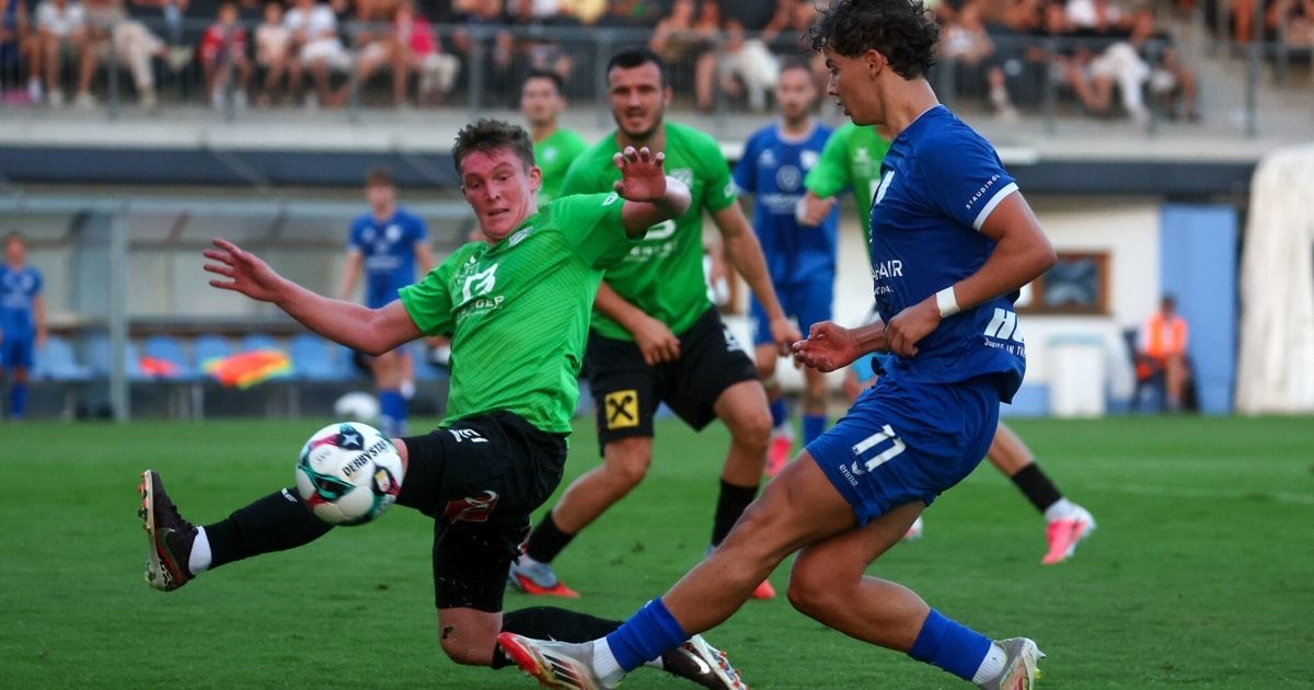 Ein Fußballspieler in einem grünen Trikot tritt den Ball, während ein anderer Spieler in einem blauen Trikot versucht, ihn zu blockieren. Sie spielen auf einem Fußballfeld mit anderen Spielern und Zuschauern im Hintergrund.