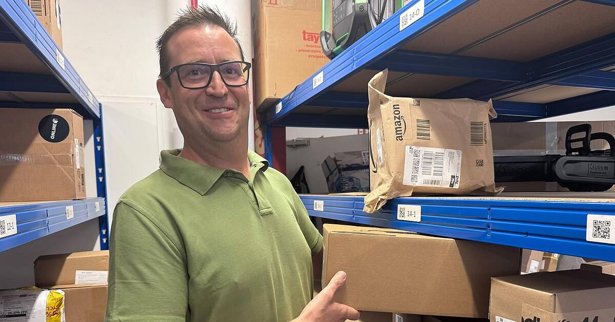 Ein Mann in einem grünen Poloshirt steht in einem Lager und zeigt auf ein braunes Paket auf einem blauen Regal.
