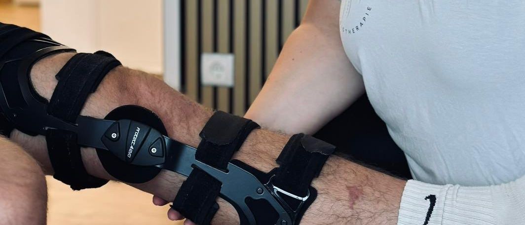 Eine Frau in einem weißen T-Shirt hilft einem Mann mit einer Knieorthese, sich in einen Stuhl zu setzen. Der Mann hat einen Verband am Knie. Sie befinden sich in einem Raum mit weißen Wänden.
