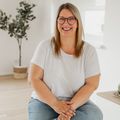 Eine Frau mit Brille sitzt auf dem Boden, lächelt und posiert für ein Foto in einem weißen Raum.