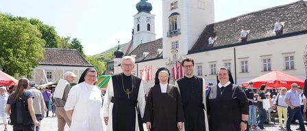 Eine Gruppe von Geistlichen und Nonnen posiert für ein Foto vor einem historischen Gebäude mit einem Turm und Fahnen im Hintergrund.