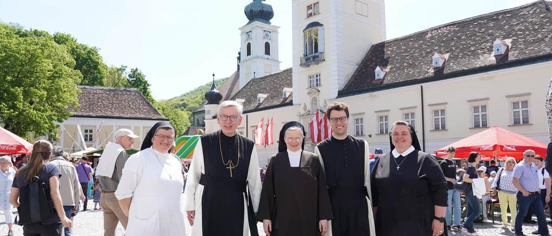 Eine Gruppe von Geistlichen und Nonnen posiert für ein Foto vor einem historischen Gebäude mit einem Turm und Fahnen im Hintergrund.