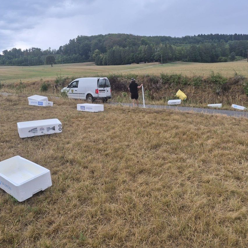 Eine Person steht in einem Feld, in dem ein weißer Van geparkt ist. Mehrere weiße Kisten sind verstreut und eine gelbe Tasche liegt auf dem Boden.
