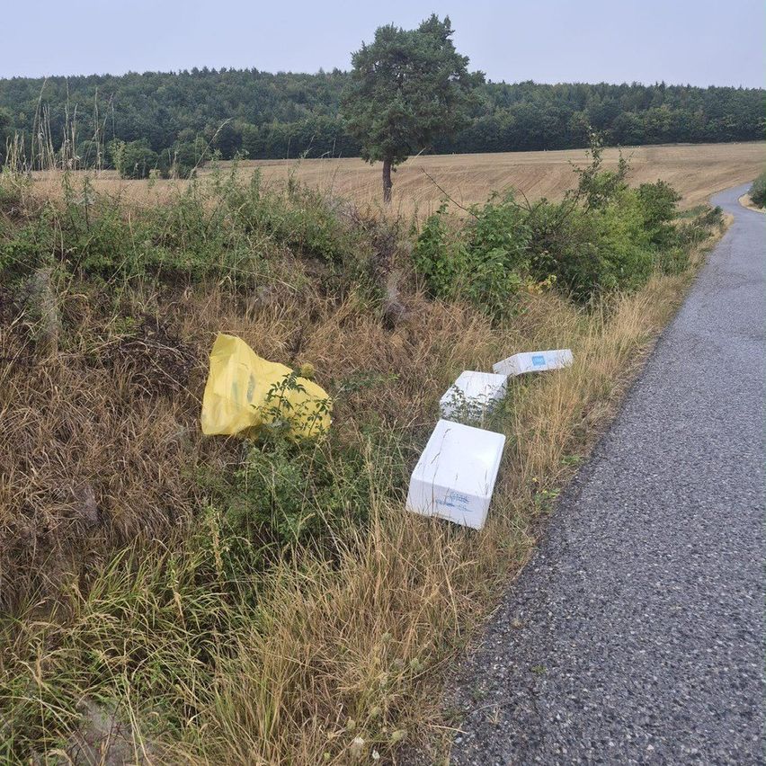 Eine ländliche Straßenszene mit einem gelben Plastikbeutel und drei weißen Kisten, die in der Nähe eines Kiesweges im Gras verstreut sind.