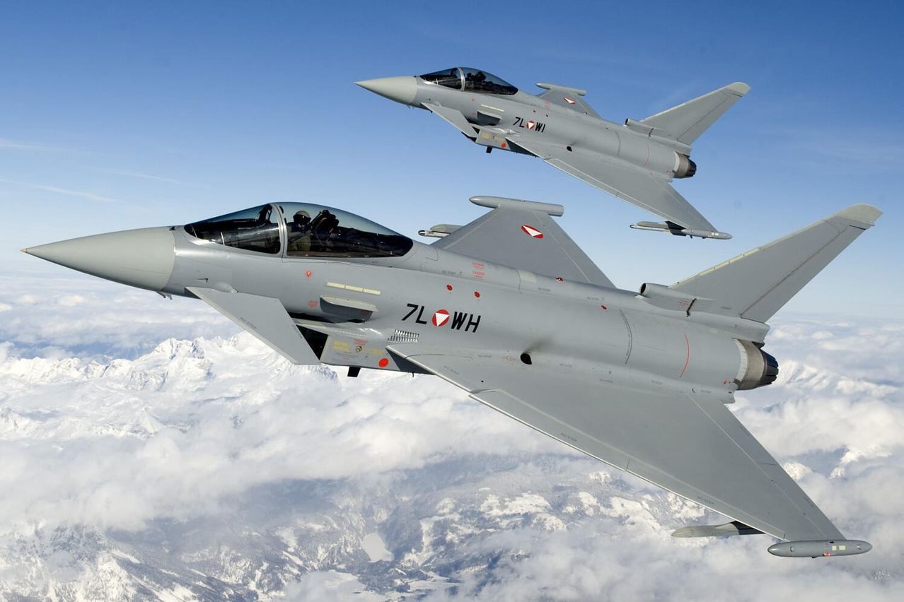 Zwei Kampfjets fliegen nebeneinander über einer verschneiten Bergkette. Einer hat '7L' und 'WH' an seiner Seite.