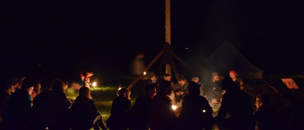 Eine Gruppe von Menschen versammelt sich bei Nacht um ein Lagerfeuer, einige mit brennenden Kerzen, unter klarem Himmel.