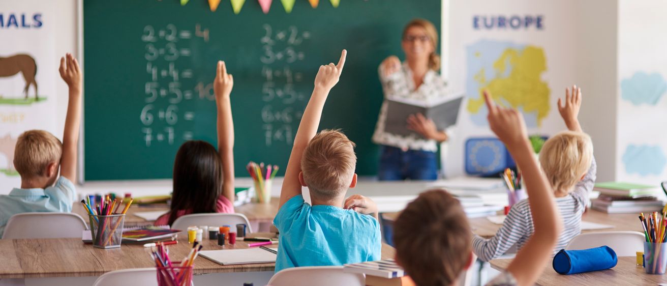 Ein Klassenzimmer mit Kindern, die ihre Hände heben, um eine Frage der Lehrerin zu beantworten. Die Lehrerin steht vor einer Tafel mit Matheaufgaben. Das Klassenzimmer ist mit bunten Wimpelketten dekoriert.
