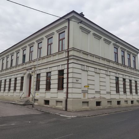 Ein großes weißes Gebäude mit vielen Fenstern und einer Treppe, die zum Eingang führt, steht an einer Straßenecke.