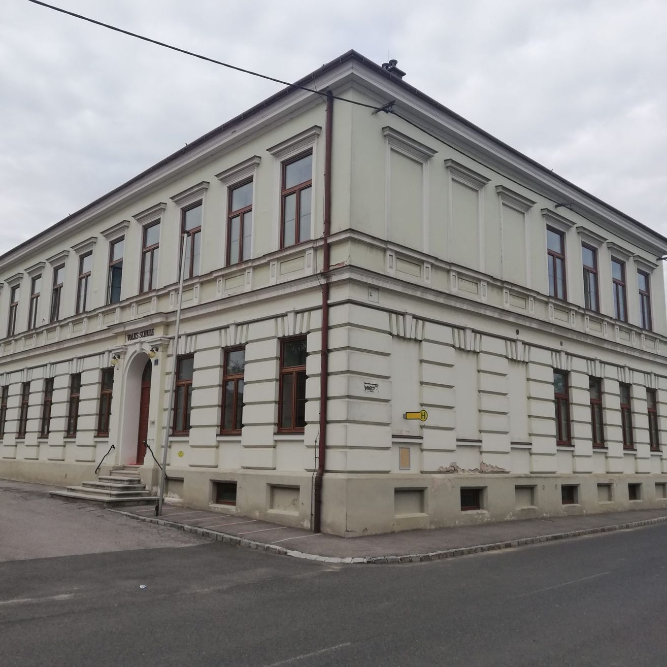 Ein großes weißes Gebäude mit vielen Fenstern und einer Treppe, die zum Eingang führt, steht an einer Straßenecke.