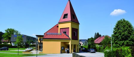 A bright yellow building with a red roof and steeple stands in a green area. A woman stands near the entrance. A car is parked nearby. Trees and other buildings are in the background.