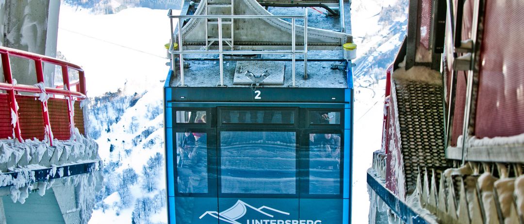 Eine blaue Seilbahn mit der Aufschrift 'Untersberg Bahn' fährt durch die verschneiten Berge. Der Wagen hat Glasfenster und ist von einem Metallgeländer umgeben.