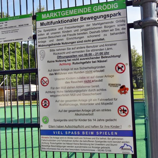 Ein Schild an einem Zaun in einem Park in Grodig, Österreich. Es sagt, dass der Park für Kinder und Jugendliche ist und bittet Besucher, rücksichtsvoll zu sein. Hunde, Müllentsorgung, Alkohol und das Fahren von Fahrzeugen sind verboten. Der Park ist von 8 Uhr bis 21 Uhr geöffnet.