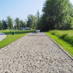 Ein mit Kies bedeckter Weg führt zu einer weißen Plattform, die von einem grünen Rasen umgeben ist. Bäume und Büsche sind im Hintergrund. Eine Straße befindet sich auf der linken Seite.