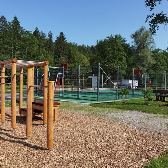 Freiluftspielplatz mit Basketballplatz und Holzgeräten, umgeben von Bäumen.