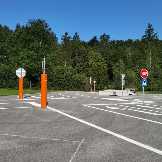 Ein leerer Parkplatz mit orangefarbenen Pfosten und weißen Markierungen. Dahinter stehen Bäume und ein Stoppschild auf der rechten Seite.