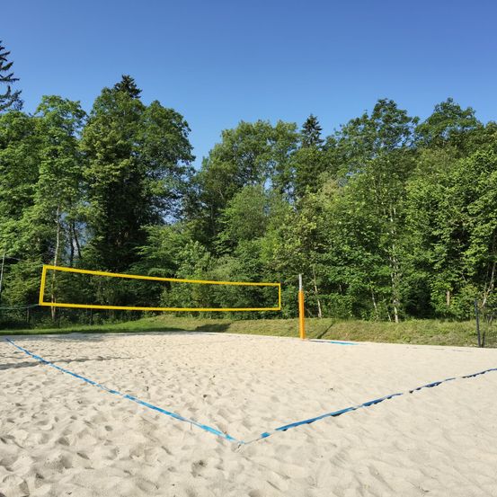 Ein Volleyballfeld im Freien mit einem gelben Netz ist von üppigen grünen Bäumen und klarem blauem Himmel umgeben. Der sandige Boden hat blaue Linien, die den Spielbereich markieren.