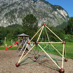 Ein Spielplatz mit Klettergeräten und einer Rutsche vor einem Bergpanorama an einem sonnigen Tag.