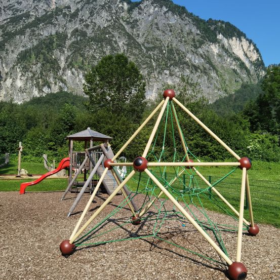 Ein Spielplatz mit Klettergeräten und einer Rutsche vor einem Bergpanorama an einem sonnigen Tag.