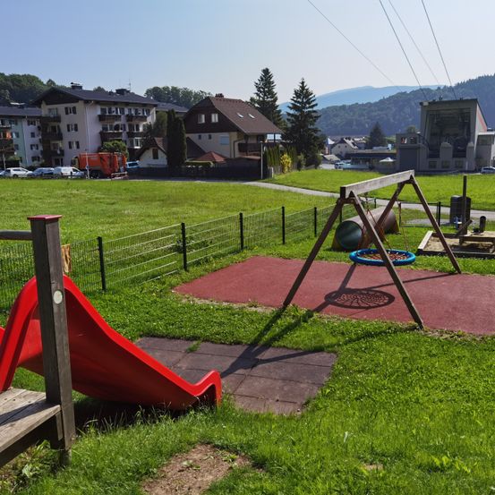 Ein Kinderspielplatz mit Rutsche, Schaukel und Häusern im Hintergrund. Der Spielplatz ist mit einem Zaun eingezäunt.