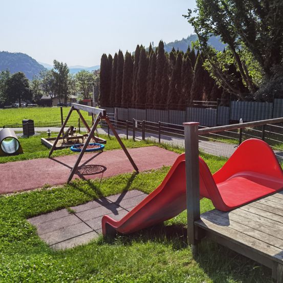 Ein Spielplatz mit einem roten Rutschbrett, einer Schaukel und einem kleinen Tunnel bei sonnigem Wetter, umgeben von grünem Gras und einem hohen Zaun.