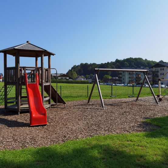 Ein Spielplatz im Freien mit einem roten Rutschvergnügen, Schaukeln und einem kleinen Unterstand. Er ist von einem Grasfeld und einem Zaun umgeben. Bäume und Gebäude sind im Hintergrund sichtbar.