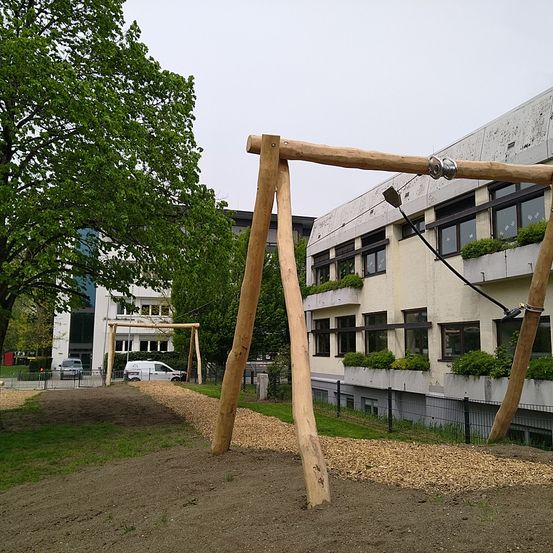 Ein Spielplatz mit hölzernen Schaukeln steht vor einem Gebäude mit vielen Fenstern und Pflanzen. Bäume und ein Weg sind in der Nähe. Ein weißer Lieferwagen ist in der Ferne geparkt.