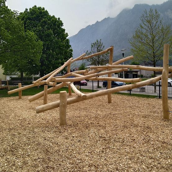 Ein Spielplatz mit hölzernen Kletterstrukturen in einem Park mit Bergen im Hintergrund und klarem Himmel.