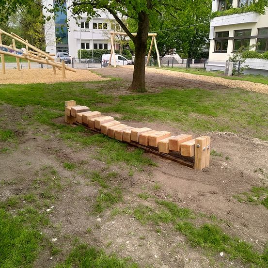 Ein Kinderspielplatz mit Holzstrukturen, einem Baum und einer Rasenfläche, umgeben von Gebäuden und geparkten Autos.