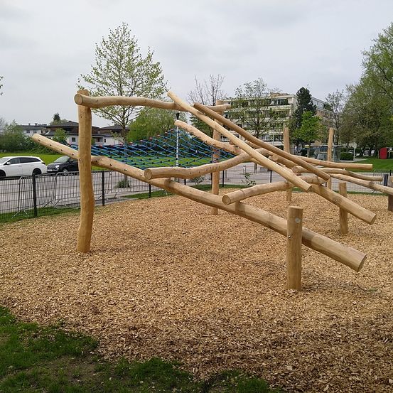 Ein hölzerner Hindernisparcours steht in einem Park mit einer Rasenfläche und einem Kiesboden. Bäume und ein Gebäude sind im Hintergrund.