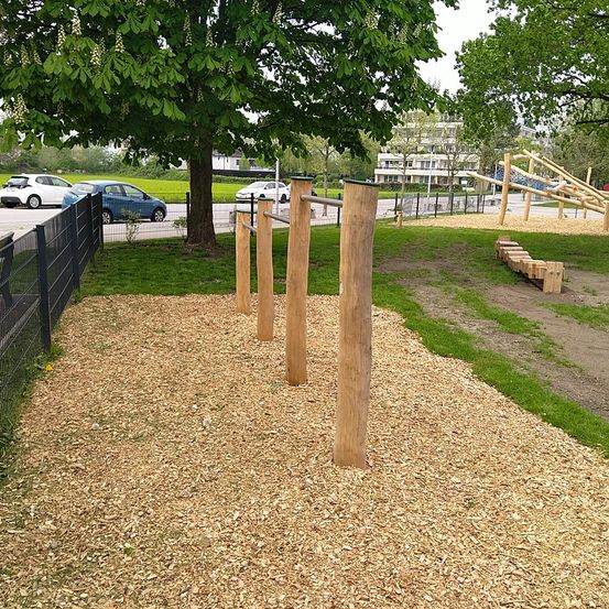 Ein Park mit einem Holzspielplatz und einem hölzernen Klettergerüst, umgeben von Gras, einem Zaun und geparkten Autos.