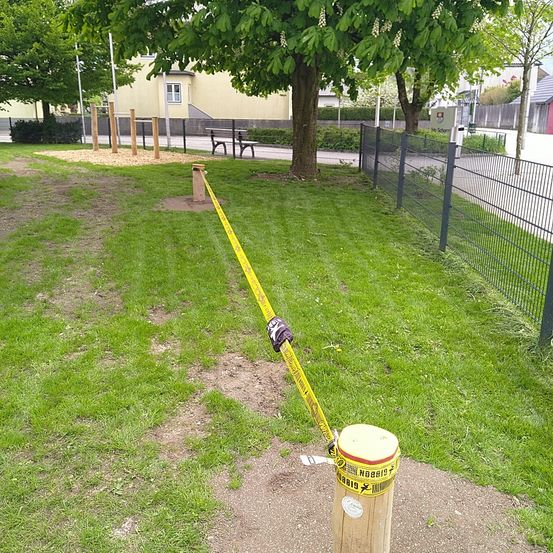 Ein gelbes Warnband ist um einen Holzpfosten in einem Park gewickelt. In der Nähe befinden sich ein Baum, eine Bank und ein Spielplatz mit Holzstangen.