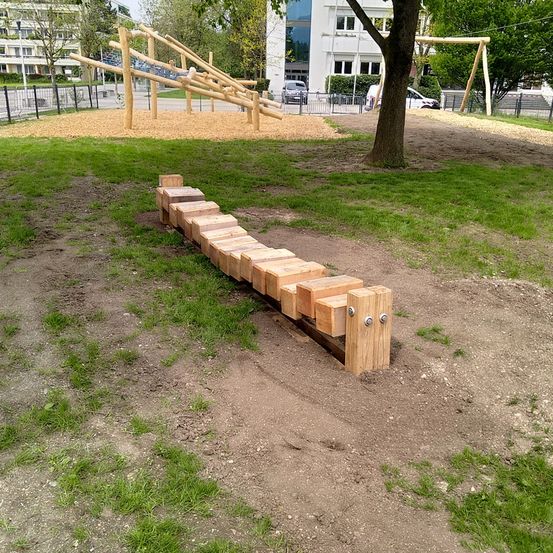 Eine Holzspielstruktur mit Rutsche und Klettergerüst befindet sich in einem Grasbereich in der Nähe eines Gebäudes. Ein Baum steht in der Nähe.