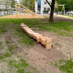 Eine Holzspielstruktur mit Rutsche und Klettergerüst befindet sich in einem Grasbereich in der Nähe eines Gebäudes. Ein Baum steht in der Nähe.