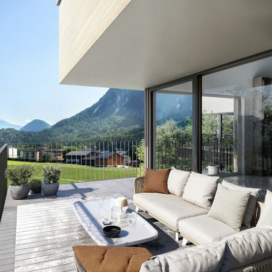 Ein Balkon mit einer Couch, einem Tisch und Pflanzen. Der Balkon hat ein Geländer und einen Blick auf ein Feld mit einem Haus und Bergen.