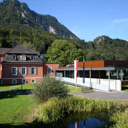 Ein modernes Gebäude mit Glaswänden steht vor einem traditionellen Backsteingebäude. Beide Strukturen sind von üppiger Vegetation und Bergen umgeben.