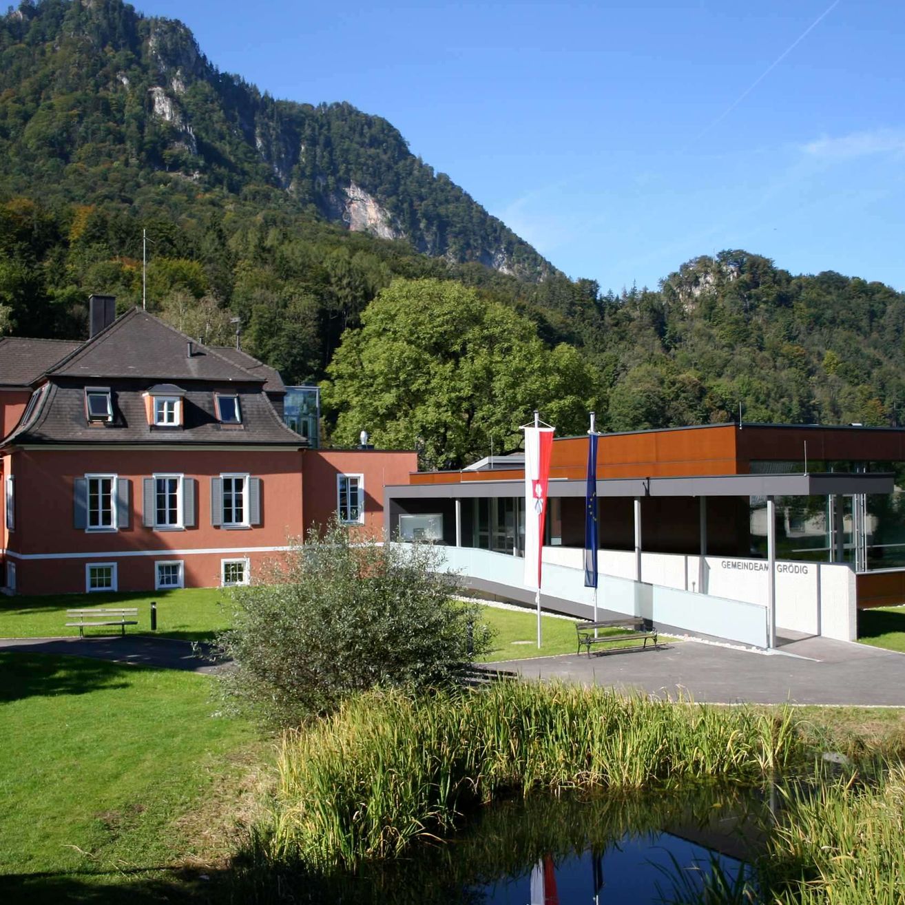 Ein modernes Gebäude mit Glaswänden steht vor einem traditionellen Backsteingebäude. Beide Strukturen sind von üppiger Vegetation und Bergen umgeben.