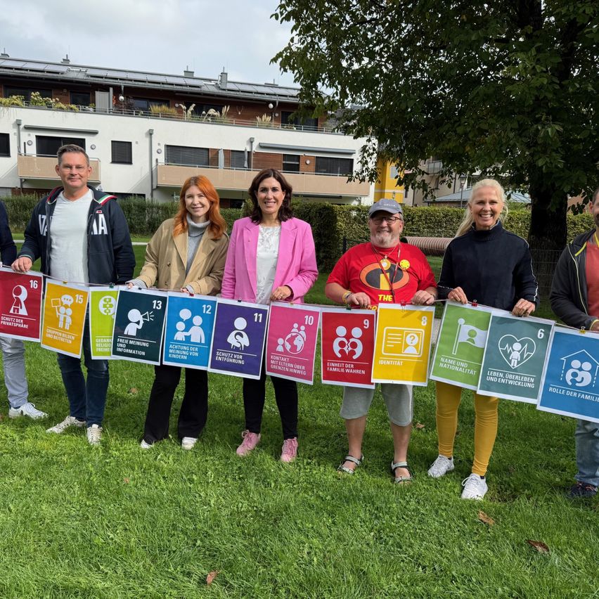 Six adults stand in a line holding a banner made of 16 colorful signs. They smile for the camera. Behind them is a building with solar panels.