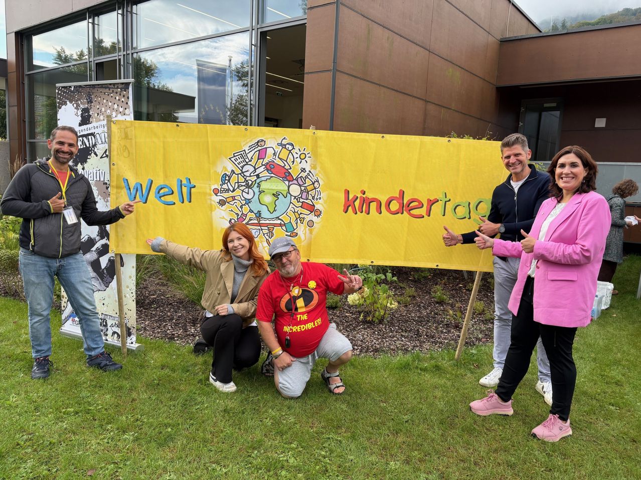 A group of adults and children pose for a photo in front of a banner with the words 'Welt kindertag' and colorful illustrations. They are smiling and appear to be posing for a picture.
