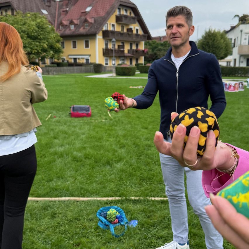A man holding multicolored balls stands on a lawn with a woman. Behind them, a house with a balcony is visible.