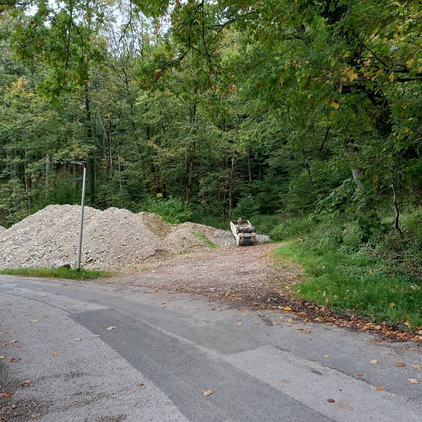 A forest road with a pile of gravel and a small dump truck. The area is surrounded by trees, with some fallen leaves on the ground.