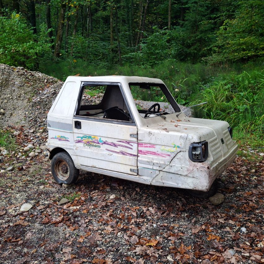 An old, white, weathered car with faded pink and blue designs sits on gravel with dried leaves. Behind it, there's a pile of rubble and a dense green forest.