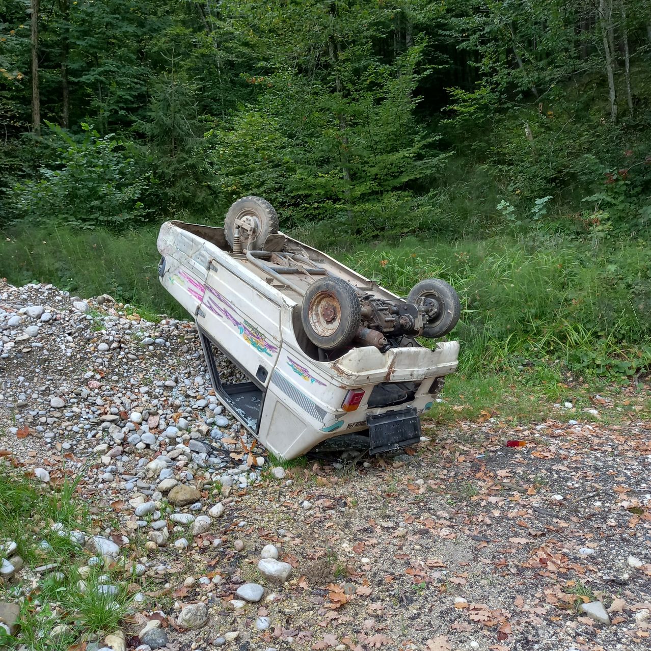An overturned RV with its tires up is on a gravel path surrounded by grass and trees.