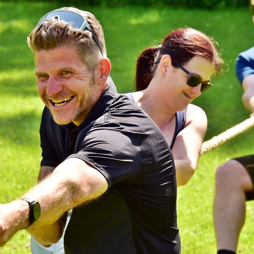 Ein Mann, der lächelt und an einem Seil zieht, mit zwei Frauen hinter ihm. Eine Frau trägt eine Sonnenbrille und hat einen Pferdeschwanz.