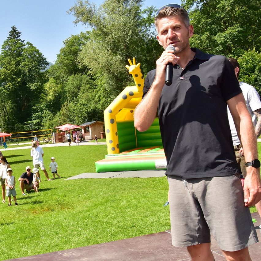 Ein Mann in einem schwarzen Poloshirt spricht in ein Mikrofon bei einer Veranstaltung im Freien mit einem bunten Hüpfburg und spielenden Kindern im Hintergrund.