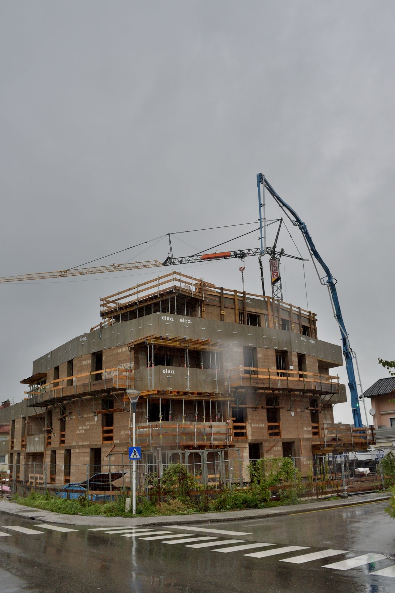 Ein laufendes Bauprojekt mit mehreren Kränen im Einsatz an einem bewölkten Tag. Das Gebäude hat mehrere Stockwerke mit Ziegelwänden, Holzgerüsten und einem aktiven Wassersprühsystem.