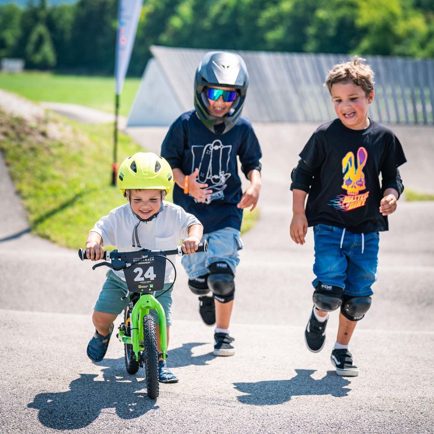 Drei Jungen fahren auf einer asphaltierten Straße mit Fahrrädern. Der Junge vorne hat einen gelben Helm und die Nummer 24 auf seinem Fahrrad. Die anderen beiden laufen hinter ihm her. Sie alle tragen Knieschützer.